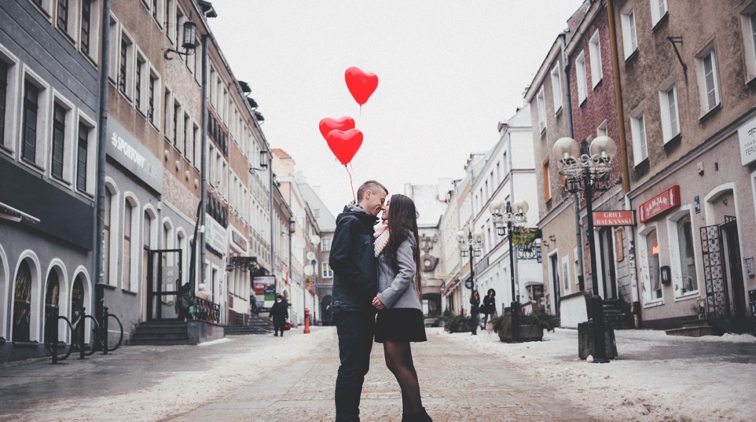 Άντρας και γυναίκα φιλιούνται κρατώντας κόκκινα μπαλόνια καρδιές