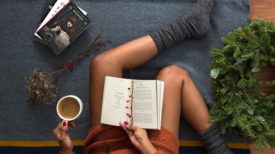 Κοπέλα καθισμένη στο πάτωμα κρατάει ανοιχτό βιβλίο και κούπα με ζεστή σοκολάτα