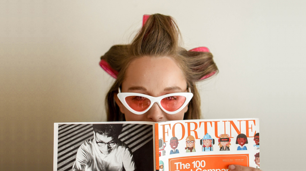 Γυναίκα με ρόλεϊ στα μαλλιά, γυαλιά ηλίου και ανοιγμένο περιοδικό μπροστά στο πρόσωπό της