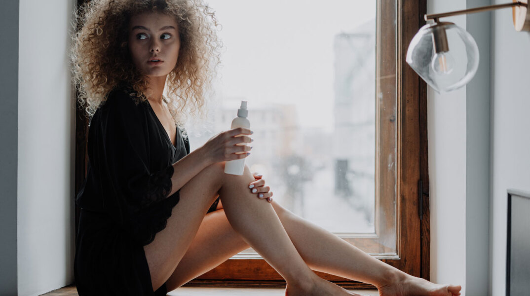 Κοπέλα με σγουρά μαλλιά καθισμένη δίπλα σε παράθυρο φορώντας μαύρα ρούχα βάζει κρέμα στα πόδια της