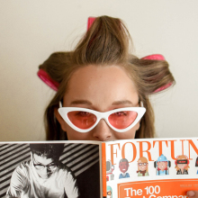 Γυναίκα με ρόλεϊ στα μαλλιά, γυαλιά ηλίου και ανοιγμένο περιοδικό μπροστά στο πρόσωπό της