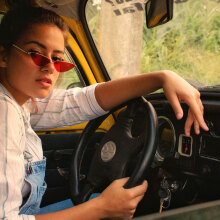 Γυναίκα καθισμένη σε αυτοκίνητο, κρατάει τιμόνι και κοιτάζει την κάμερα