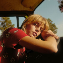 Κοπέλα με κόκκινη μπλούζα με λουλούφια καθισμένη σε αυτοκίνητο ακουμπάει τα χέρια και το κεφάλι της στο τιμόνι ενώ κοιτάζει την κάμερα