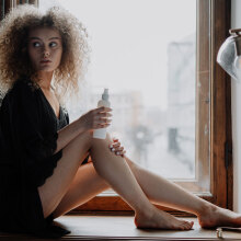 Κοπέλα με σγουρά μαλλιά καθισμένη δίπλα σε παράθυρο φορώντας μαύρα ρούχα βάζει κρέμα στα πόδια της