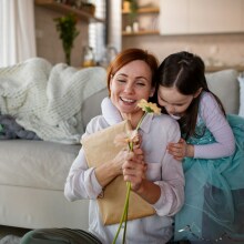 Μητέρα κρατά στα χέρια της λουλούδια και ένα πακέτο όσο την αγκαλιάζει η κόρη της. Στα αριστερά της στέκεται ακόμη ένα παιδί