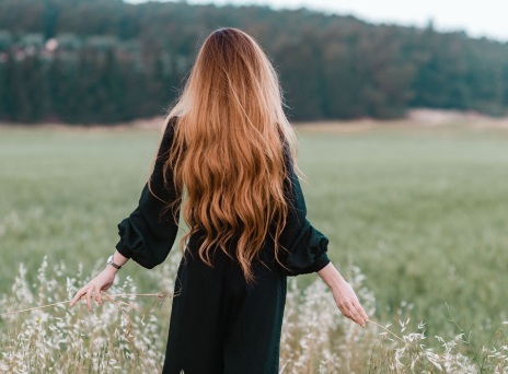 Κοπέλα με μακριά μαλλιά και γυρισμένη πλάτη περπατάει σε χωράφι αγγίζοντας τα φυτά