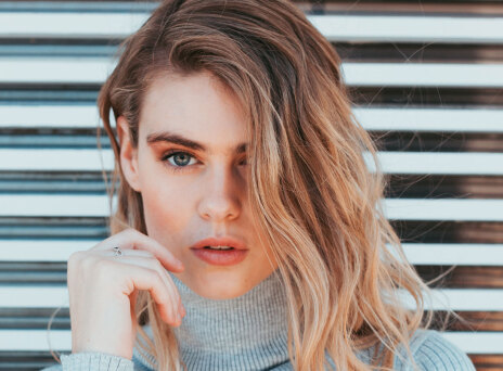 Ξανθιά κοπέλα με γκρι πλεκτό ζιβάγκο και φόντο ξύλινο φράκτη