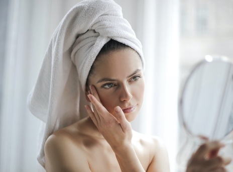 Γυναίκα με πετσέτα στο κεφάλι, με το ένα χέρι κρατάει καθρέφτη και με το άλλο αγγίζει το πρόσωπό της