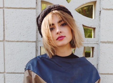 Ξανθιά κοπέλα με φούτερ και μπερέ ποζάρει μπροστά από λευκό τοίχο με σκαλιστό παραθυράκι