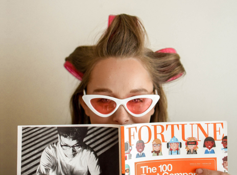 Γυναίκα με ρόλεϊ στα μαλλιά, γυαλιά ηλίου και ανοιγμένο περιοδικό μπροστά στο πρόσωπό της