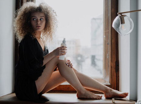Κοπέλα με σγουρά μαλλιά καθισμένη δίπλα σε παράθυρο φορώντας μαύρα ρούχα βάζει κρέμα στα πόδια της