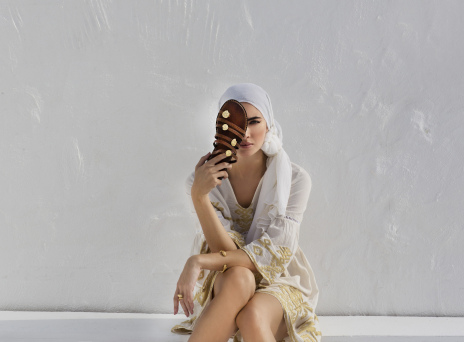 Γυναίκα καθισμένη σε πεζούλι με λευκά ρούχα κρατά μπροστά στο πρόσωπό της σανδάλι