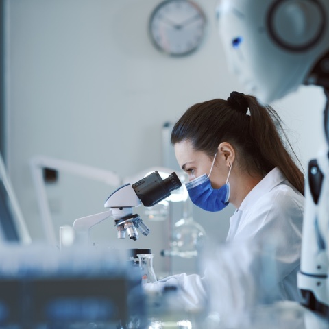 Γυναίκα με μάσκα που κοιτά σε μικροσκόπιο και δίπλα της ένα ρομπότ