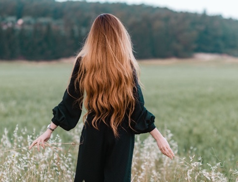 Κοπέλα με μακριά μαλλιά και γυρισμένη πλάτη περπατάει σε χωράφι αγγίζοντας τα φυτά