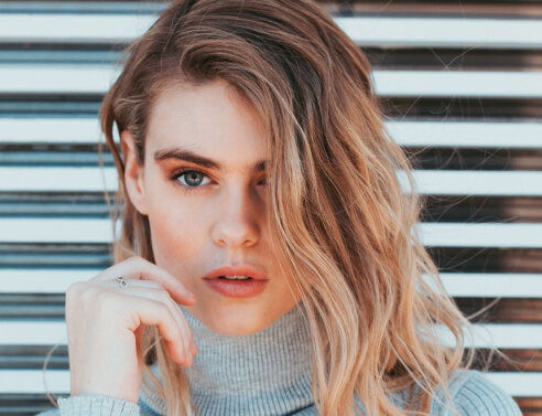 Ξανθιά κοπέλα με γκρι πλεκτό ζιβάγκο και φόντο ξύλινο φράκτη