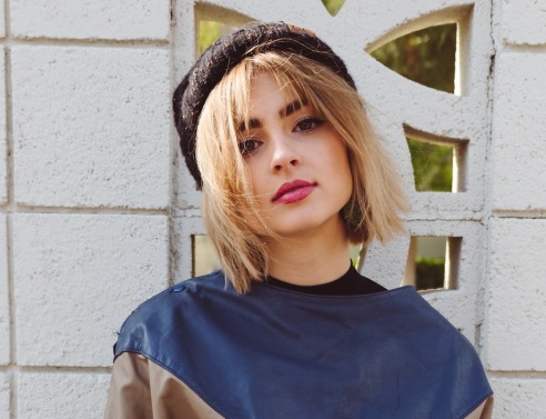 Ξανθιά κοπέλα με φούτερ και μπερέ ποζάρει μπροστά από λευκό τοίχο με σκαλιστό παραθυράκι