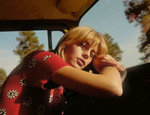 Κοπέλα με κόκκινη μπλούζα με λουλούφια καθισμένη σε αυτοκίνητο ακουμπάει τα χέρια και το κεφάλι της στο τιμόνι ενώ κοιτάζει την κάμερα