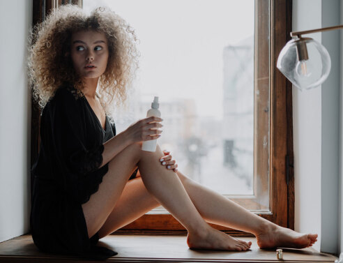 Κοπέλα με σγουρά μαλλιά καθισμένη δίπλα σε παράθυρο φορώντας μαύρα ρούχα βάζει κρέμα στα πόδια της