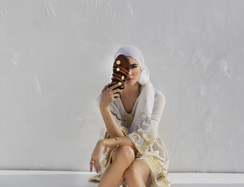 Γυναίκα καθισμένη σε πεζούλι με λευκά ρούχα κρατά μπροστά στο πρόσωπό της σανδάλι