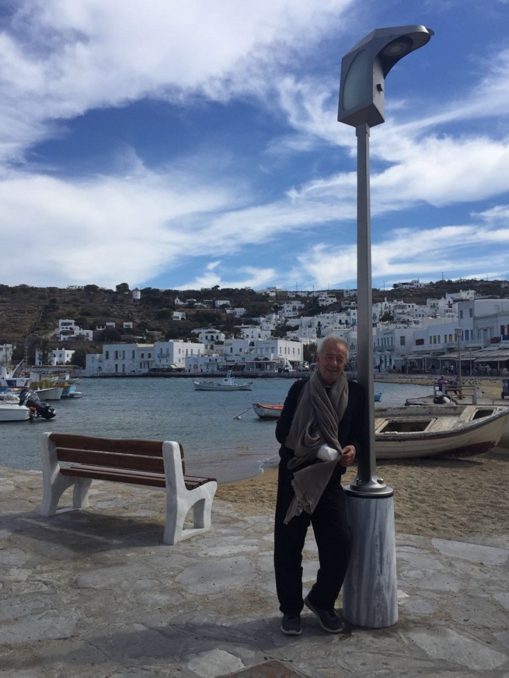 Ο σχεδιαστής Μηνάς στέκεται ανάμεσα στο παγκάκι και τη λάμπα δρόμου που σχεδίασε για τη Μύκονο