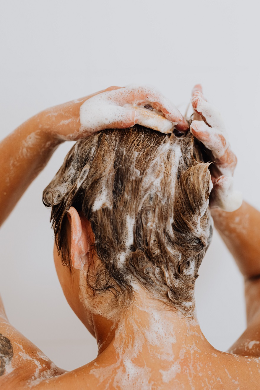 Κοπέλα με γυρισμένη πλάτη κάνει μασάζ στα μαλλιά της με σαμπουάν που έχει αφρίσει